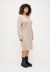 Beige kötött ruha hosszú ujjal, V-nyakkivágással, félhosszú fazonnal, fekete bokacsizmával, vastag talppal párosítva.