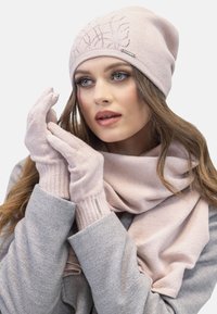 Berretto a maglia rosa chiaro con design in rilievo, guanti coordinati e una morbida sciarpa. Il tessuto è testurizzato, accogliente e leggero. Indumento esterno grigio visibile.