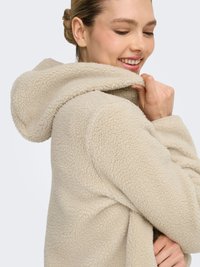 Beige fleece jacket with a hood, featuring a textured surface and a loose fit. The model holds the jacket close to the neck.