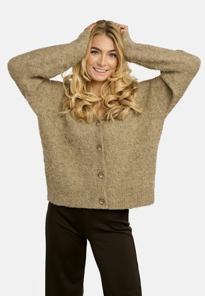 Donna bionda sorridente con le mani sulla testa, indossa un cardigan beige morbido con bottoni e pantaloni neri su uno sfondo semplice.