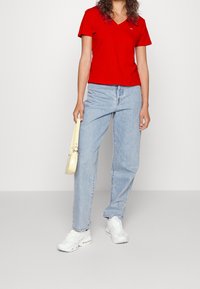 T-shirt rossa a scollo a V abbinata a jeans blu chiaro a gamba dritta. Gli accessori includono una piccola borsa gialla e sneakers bianche.