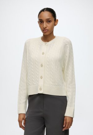 Frau trägt eine cremefarbene, kabelgestrickte, geknöpfte Strickjacke mit langen Ärmeln und dunkelgraue Hose, steht vor einem einfarbigen weißen Hintergrund.