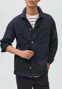 Čierna džínová bunda so skrytými náprsními vreckami, gombíkovým zapínaním a prehnutým golierom. Noste ju cez pruhovanú košeľu, ktorá má voľný strih.