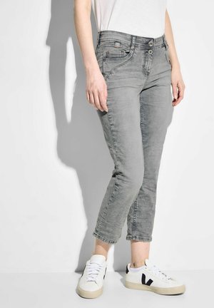 Personne portant un jean gris clair court et des baskets blanches avec un logo noir "V", debout contre un mur blanc uni et un sol blanc.