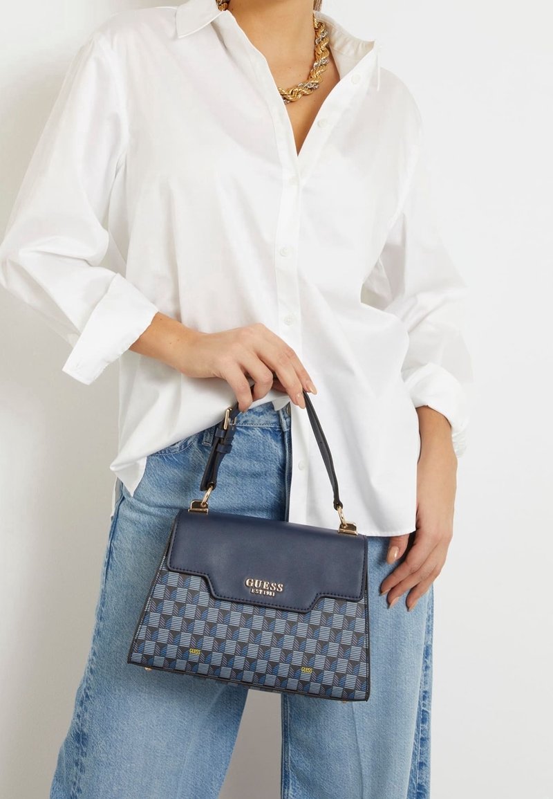 Personne portant une chemise blanche boutonnée et un jean bleu, tenant un sac à main Guess bleu à motifs avec une poignée noire et des accents dorés.
