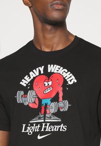 Czarny bawełniany t-shirt z nadrukiem kreskówkowego czerwonego serca podnoszącego ciężary, w niebieskich szortach i kroplami potu. Napis brzmi "CIĘŻKIE CIĘŻARY" i "LEKKIE SERCA".