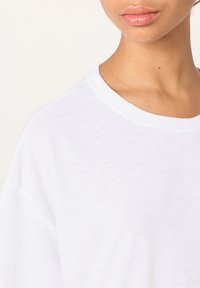 Maglietta bianca in cotone con scollo rotondo, maniche corte e tessuto leggermente strutturato. Dettaglio su spalla e area del collo.