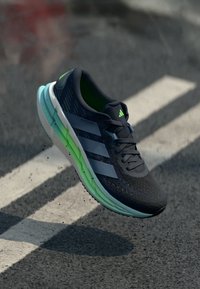 Black running shoe with green accents, mesh upper, three white stripes, textured surface, and cushioned sole. Levitating above pavement.