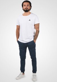Vit bomull t-shirt med en svart triangel-logotyp, tillsammans med mörkblå joggerbyxor med fickor och dragsko, samt vita sneakers.