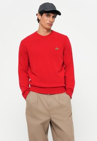 Pulover tricotat roșu, cu guler rotund, manșete și tiv cu bare. Prezintă un mic logo verde de crocodil pe pieptul stâng. Fit casual.