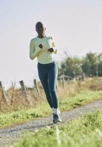 Conjunto deportivo: camiseta de manga larga amarillo claro y leggings azul oscuro. Zapatillas de running con detalles. Camino exterior rodeado de hierba y una valla de madera.