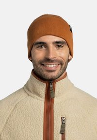 Gorro de punto acanalado naranja con una pequeña etiqueta, usado con una chaqueta de forro polar color crema que presenta un cierre con cremallera y una tela texturizada.