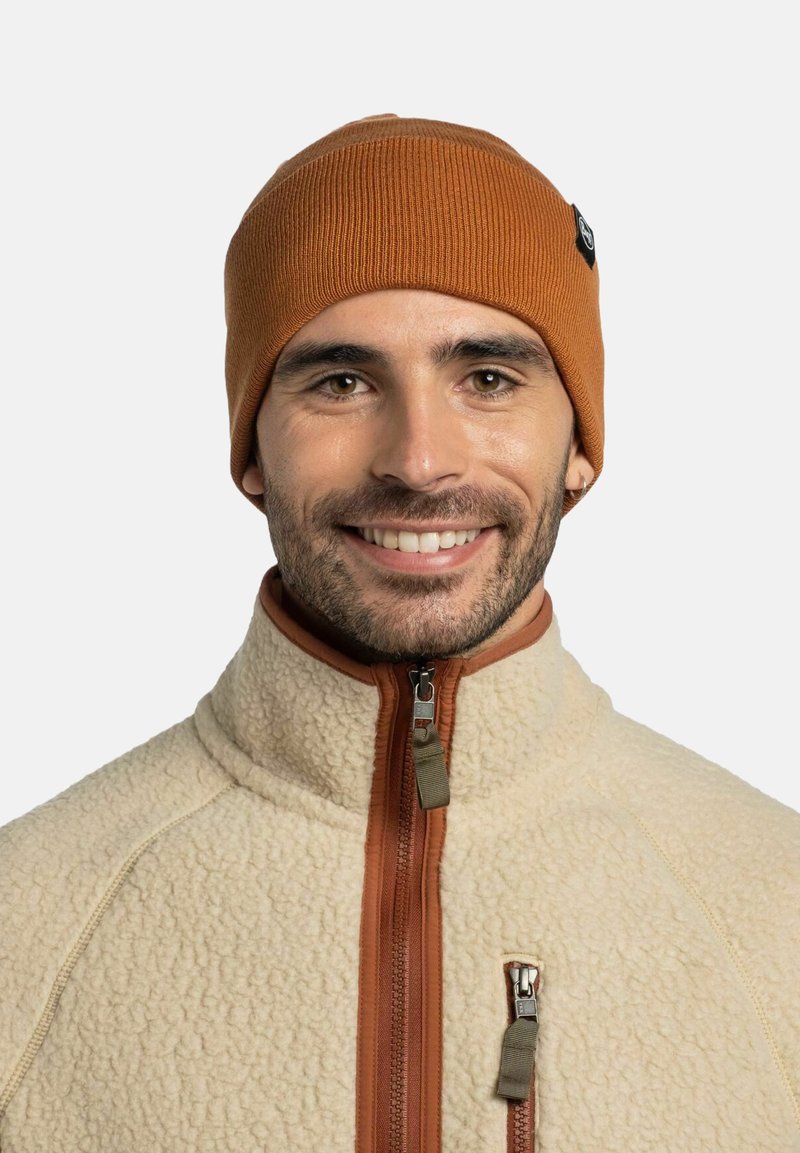 Gorro de punto acanalado naranja con una pequeña etiqueta, usado con una chaqueta de forro polar color crema que presenta un cierre con cremallera y una tela texturizada.