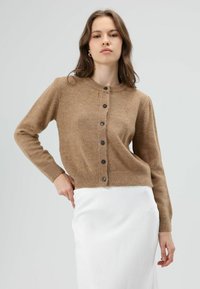 Bruine gebreide cardigan met een ronde halslijn, knoopsluiting en geribde zoom. Gecombineerd met een witte rok, met nadruk op textuur en kleurcontrast.