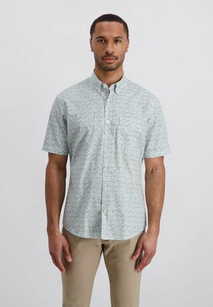 Mann trägt ein helles, kurzärmeliges Hemd mit kleinem Blumenmuster und beige Hose, steht vor einem schlichten grauen Hintergrund.