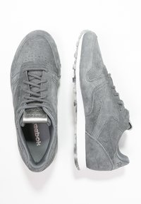 Baskets en nubuck gris de Reebok vues de dessus et de côté, avec des lacets, des semelles blanches et des logos de marque subtils sur la languette et le talon.