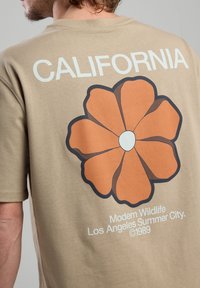 Vaaleanruskea puuvillapaita, jossa on suuri oranssi kukkakuva selkäpuolella, sanat "CALIFORNIA" yläpuolella ja vaaleansinistä tekstiä alla.