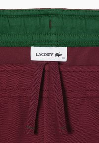 Burgunderfarbene Shorts mit einem elastischen grünen Bund, ausgestattet mit einem Kordelzug und einem weißen Etikett mit "LACOSTE" und Größe 36. Strukturiertes Gewebe.