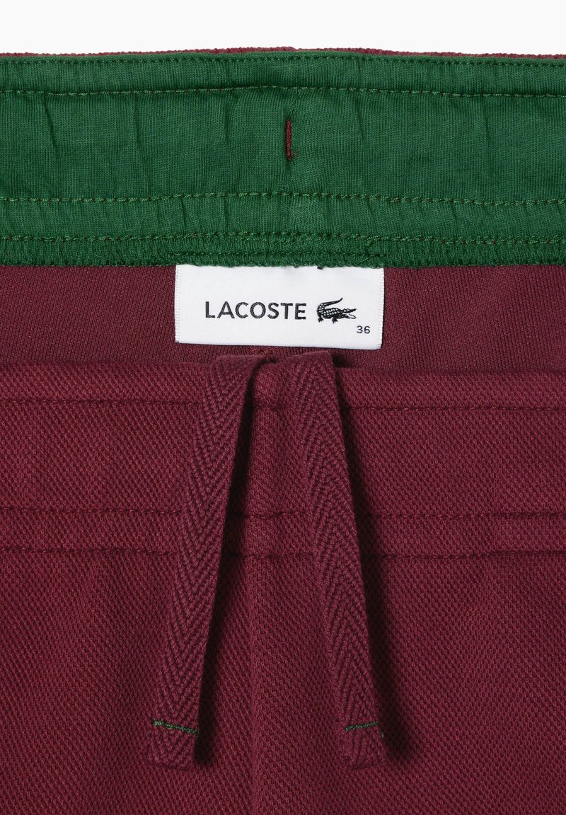 Pantaloni corti bordeaux con una cintura elastica verde, dotati di un cordoncino e un'etichetta bianca con "LACOSTE" e taglia 36. Tessuto testurizzato.