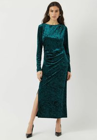 Tyrkysové sametové maxi šaty s dlouhými rukávy, bočním rozparkem a tečkovaným vzorem. Hladká textura s přiléhavou siluetou a kulatým výstřihem.
