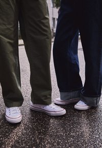 Baskets en toile blanche avec des accents rouges, associées à un pantalon cargo vert et un jean bleu foncé retroussé aux chevilles, sur une surface asphaltée texturée.