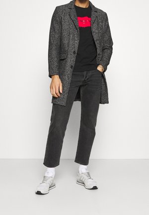 Szary płaszcz w jodełkę, czarna koszulka z czerwoną grafiką, czarne dżinsy i białe buty sportowe z szarymi akcentami. Swobodny styl, widok pełnej długości.