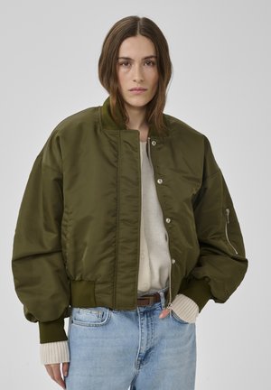 Frau trägt eine übergroße olivgrüne Bomberjacke über einem cremefarbenen Pullover und hellblauen Jeans vor einfachem grauem Hintergrund.