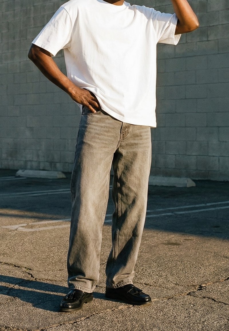Personne portant un t-shirt blanc, un jean gris ample et des chaussures noires, debout sur un trottoir fissuré, une main sur la hanche et l'autre levée vers la tête.