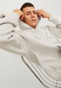 Haut à capuche gris clair avec une fermeture éclair à l'avant, une capuche et des poignets côtelés. Texture lisse, présentant une coupe ample et des détails de surpiqûre audacieux.