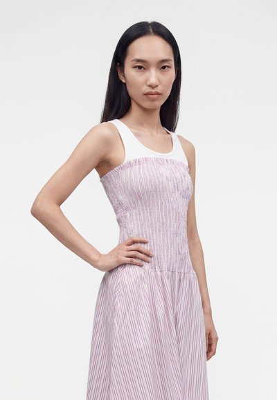 Γυναίκα με μακριά μαύρα μαλλιά που φορά αμάνικη λευκή μπλούζα και ανοιχτό ροζ φόρεμα με κάθετες ρίγες, στέκεται με το χέρι στη μέση.