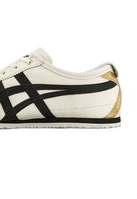 White leather sneaker with black accents and striped design. Features gold eyelets and a textured, tan-colored back detail. Rubber sole.