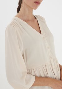 Blusa beige chiaro con scollatura a V, chiusura frontale con bottoni, vita arricciata e maniche a tre quarti realizzata in tessuto morbido e fluido.