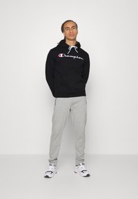 Felpa nera con cappuccio e logo "Champion", abbinata a pantaloni della tuta grigi chiari e sneaker bianche con dettagli neri.