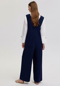 Tums zila kombinezona ar platām kājām, kabatām un pieguļošu topu. Kopā ar baltu kreklu ar pogām pie aprocēm un plakanu krēmbalts kurpēm.