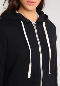 Felpa nera con zip realizzata in tessuto morbido, dotata di cappuccio con lacci color crema e tasca frontale. Design semplice e funzionale.