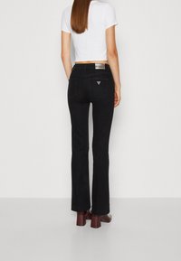 Vaqueros negros de corte acampanado con una textura suave, que cuentan con bolsillos traseros y un detalle de logo plateado en la cintura. Combinados con un top corto blanco.