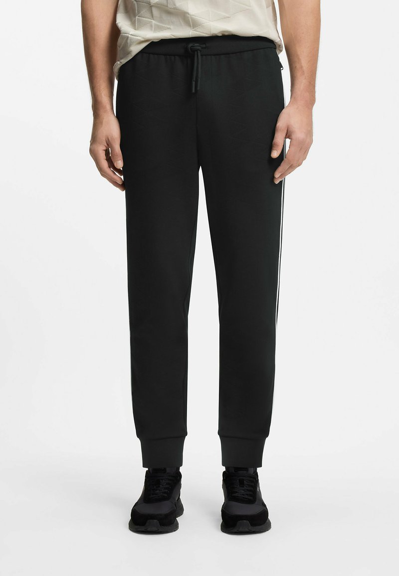 Homme portant un pantalon de jogging noir avec des poignets élastiques, une taille avec cordon, des bandes blanches sur les côtés, des baskets noires et une chemise claire à motifs.