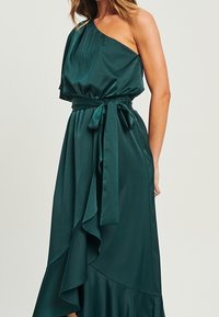 Vestido de satén esmeralda de un solo hombro con una silueta fluida, dobladillo con volantes y detalle de cintura anudada. Textura suave con un acabado brillante.