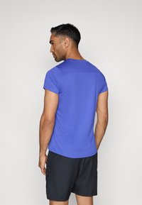 Τιράντα κοντομάνικη, εφαρμοστή αθλητική μπλούζα σε έντονο μωβ ύφασμα με ομαλή υφή, συνδυασμένη με μαύρα σορτς, απεικονισμένη από πίσω.