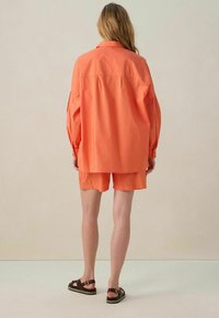 Chemise orange à boutons avec épaules larges et manches longues, associée à un short assorti. Porté avec des sandales marron foncé sur un fond neutre.