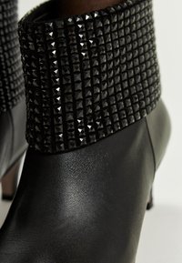 Botas de cuero negro hasta el tobillo con tacones de altura media y puños anchos decorados con remaches cuadrados negros en filas.