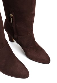 Para ciemno-brązowych zamszowych botków do połowy łydki z grubymi obcasami i złotymi bocznymi zamkami na tle białym.