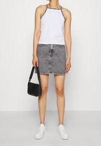 Top bianco senza maniche con bordi neri, abbinato a una gonna di jeans grigia. La modella porta una piccola borsa nera con texture e indossa sneakers bianche.