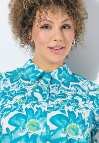 Camicia button-up con un motivo floreale in tonalità di turchese e verde su uno sfondo bianco. Tessuto morbido con colletto e bottoni.