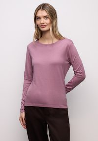 Camiseta de manga larga en tejido suave y elástico, con escote redondo y color lavanda apagado. Combinada con pantalones oscuros.