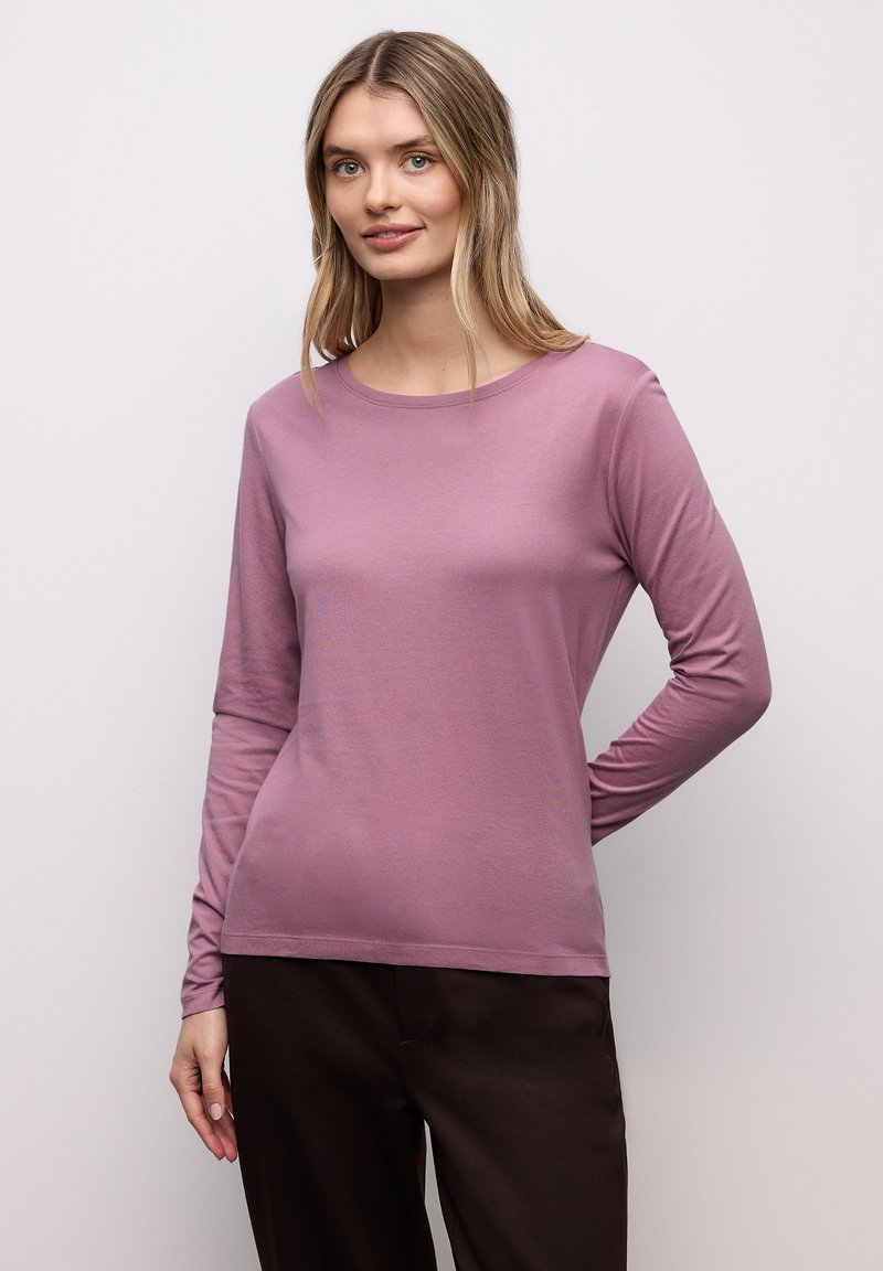 Camiseta de manga larga en tejido suave y elástico, con escote redondo y color lavanda apagado. Combinada con pantalones oscuros.