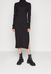 Svart långärmad klänning med hög hals, med gröna zigzagdetaljer på ärmarna och en sidoklyfta. Bärs med kraftiga svarta stövlar.