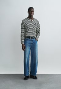 Férfi fekete-fehér csíkos hosszú ujjú ingben, kék bő szárú farmerrel, fekete övvel és fekete cipővel, egy sima világos fal előtt állva.