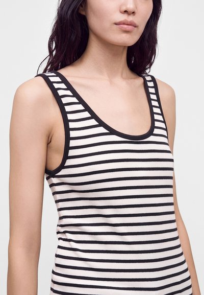 Femme portant un débardeur côtelé sans manches à rayures horizontales noires et blanches avec une bordure noire sur un fond clair.