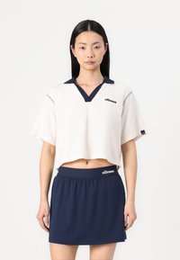 Krämfärgad kortärmad polo med en marinblå krage, prydd med en logotyp och randig trim. Matchad med en marinblå kjol med elastiskt midjeband. Slät textur.